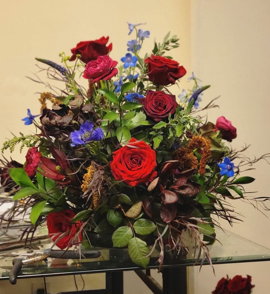 Mixed bouquet with red roses, blue delphiniums, and textured seed pods.